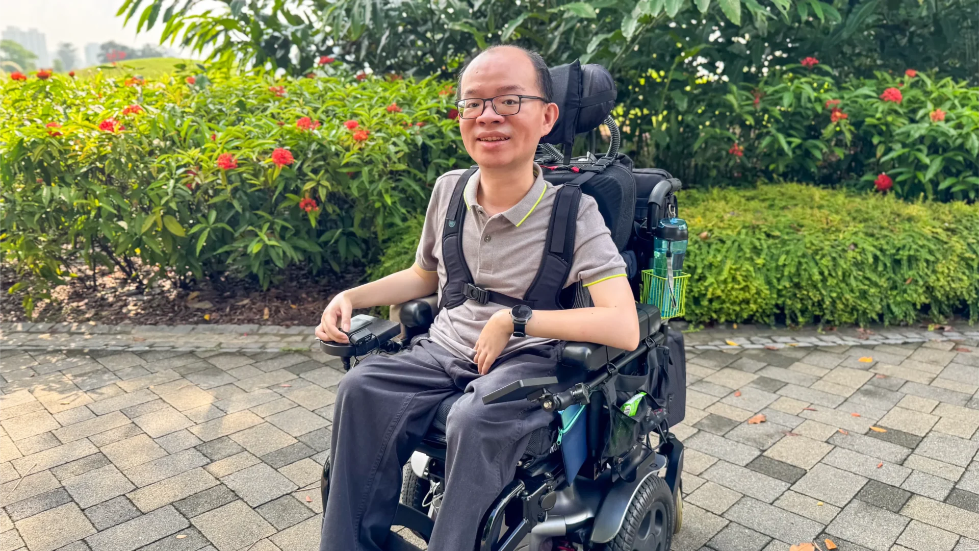 Boon Keng, smiling while sitting in his electric wheelchair. He is wearing a grey polo shirt, seated at a public park.