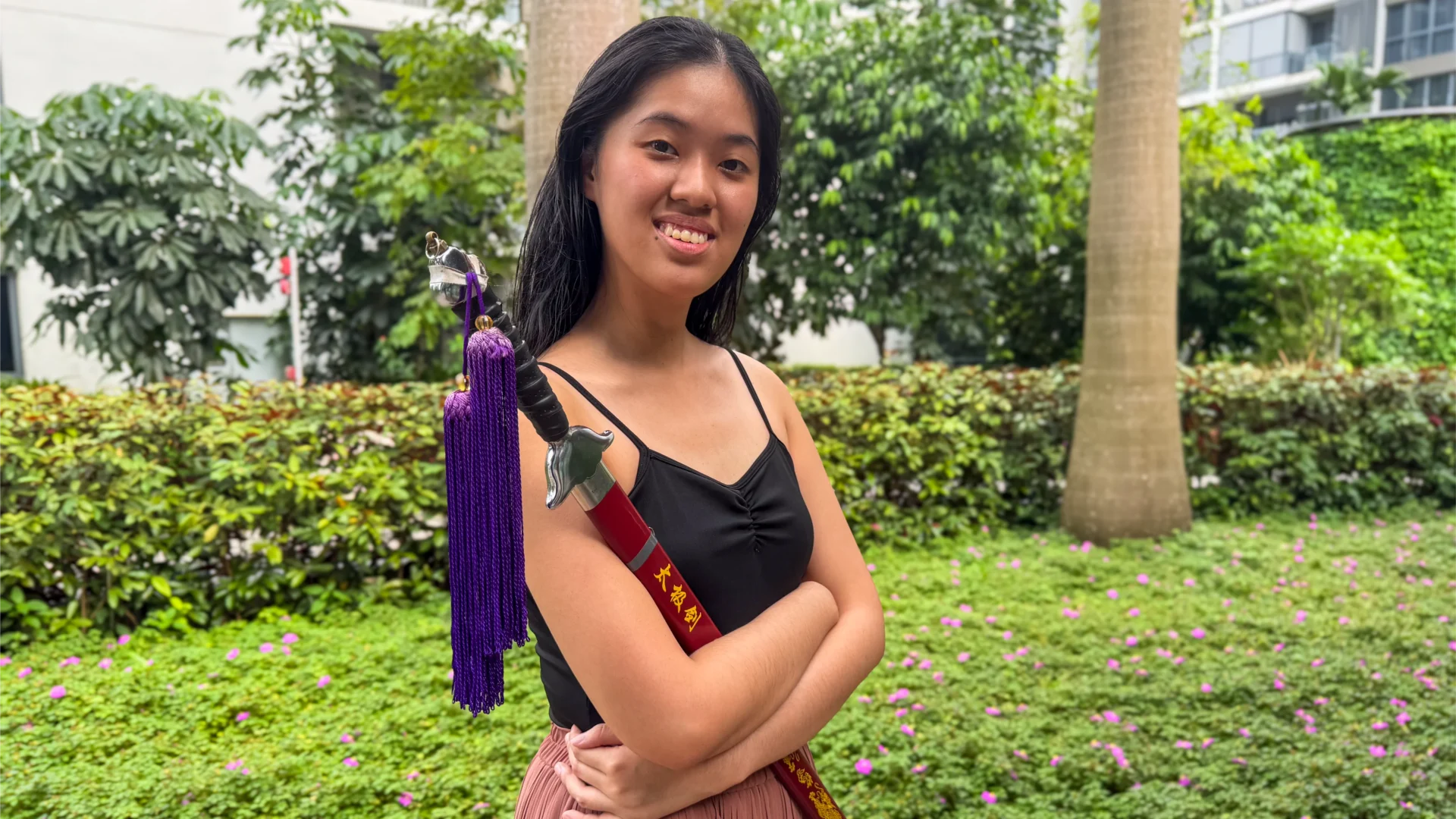 Jade wearing a bright smile on her face. She is dressed in a black leotard while holding up a sword in its sheath, up to her chest. Two purple tassels hang from the hilt of the sword.