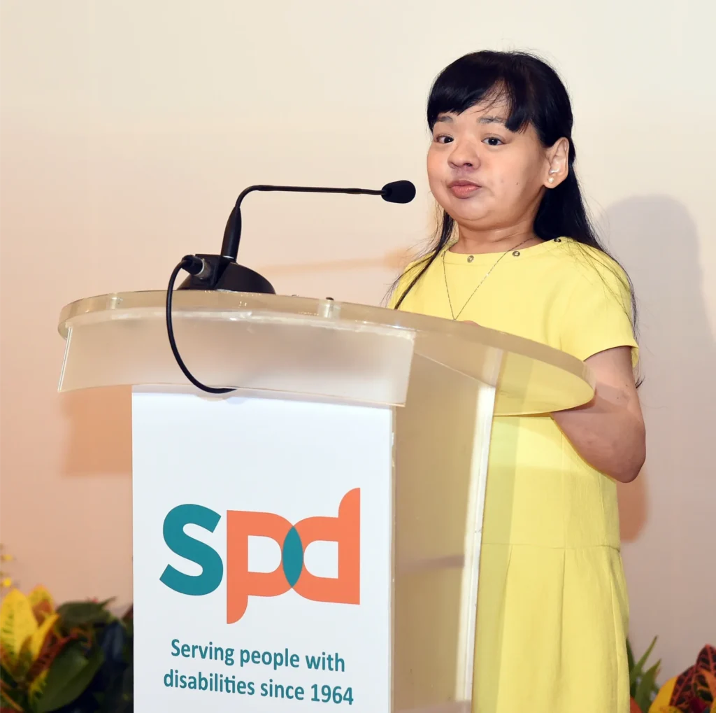 Pearl speaking on-stage, standing behind a podium and wearing a sunny yellow dress. The event is for SPD Singapore, with a sign on the podium showing the SPD logo and the text: Serving people with disabilities since 1964.
