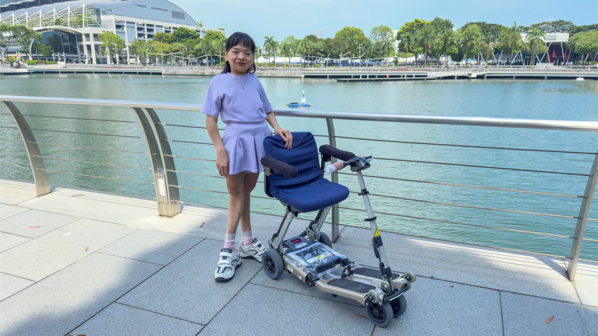 Pearl stands beside her electric scooter, dressed in a lavender skirt. The Marina Bay Sands can be seen in the background.