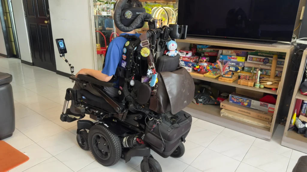 Timothy showing off the back of his current wheelchair, which is of a traditional design. There are various tubes and wires to power his wheelchair and BiPAP breathing machine. Timothy has also decorated his wheelchair with a number of pins and keychains, along with a brown leather bag and a cup holder.