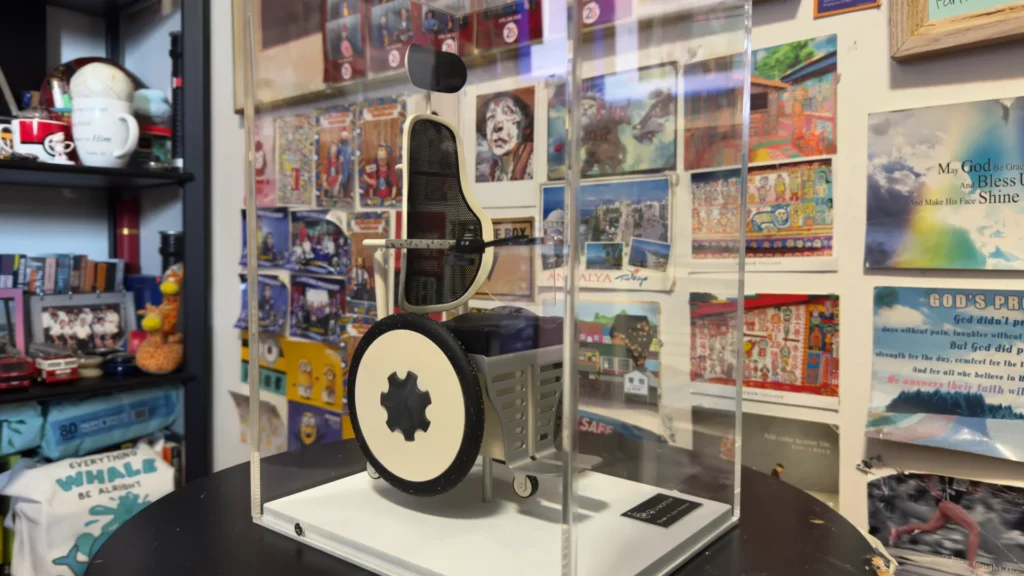 A scale model of Timothy’s dream wheelchair that he designed, displayed in a clear acrylic box. It is a re-imagining of wheelchair designs; unlike traditional wheelchairs, this model depicts features including a mesh backrest, and black and white wheels that have no spokes.
