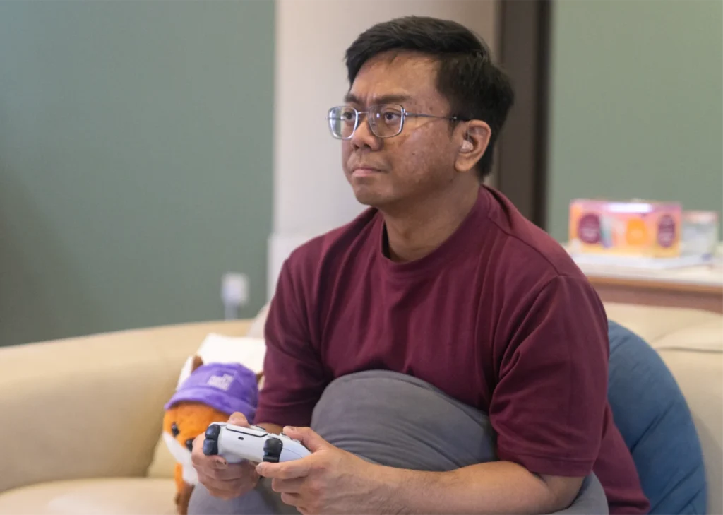Winston sits on the sofa with a game controller in his hand, a serious expression on his face as he plays video games. The fox plushie by The Purple Parade can be seen sitting behind him.