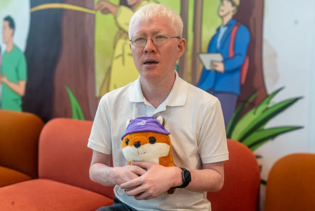 YP sitting on the sofa, holding the Purple Parade fox plushie wearing a purple bucket hat. The hat has a logo that says “The Purple Parade”. 
