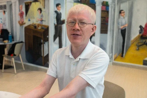 YP wearing glasses and a white polo shirt, sitting indoors with a softly blurred background of a painting and furniture. He has a pale complexion, blue-grey eyes, and light-coloured hair, with a slight smile on his face.
