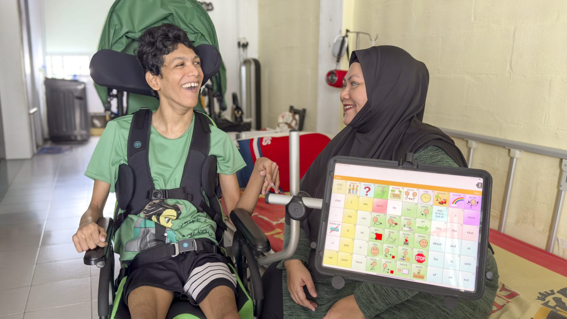 Fadillah and Sayfullah are sitting side by side, grinning widely at each other. The TouchChat app is open on Sayfullah’s iPad