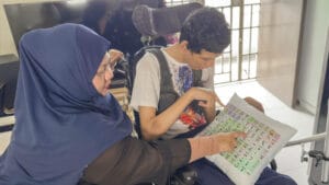 Fadillah and Sayfullah pointing at a cushion with an AAC menu printed on it. Various emotions and actions are printed on it, e.g. “Eat” and “Happy”.