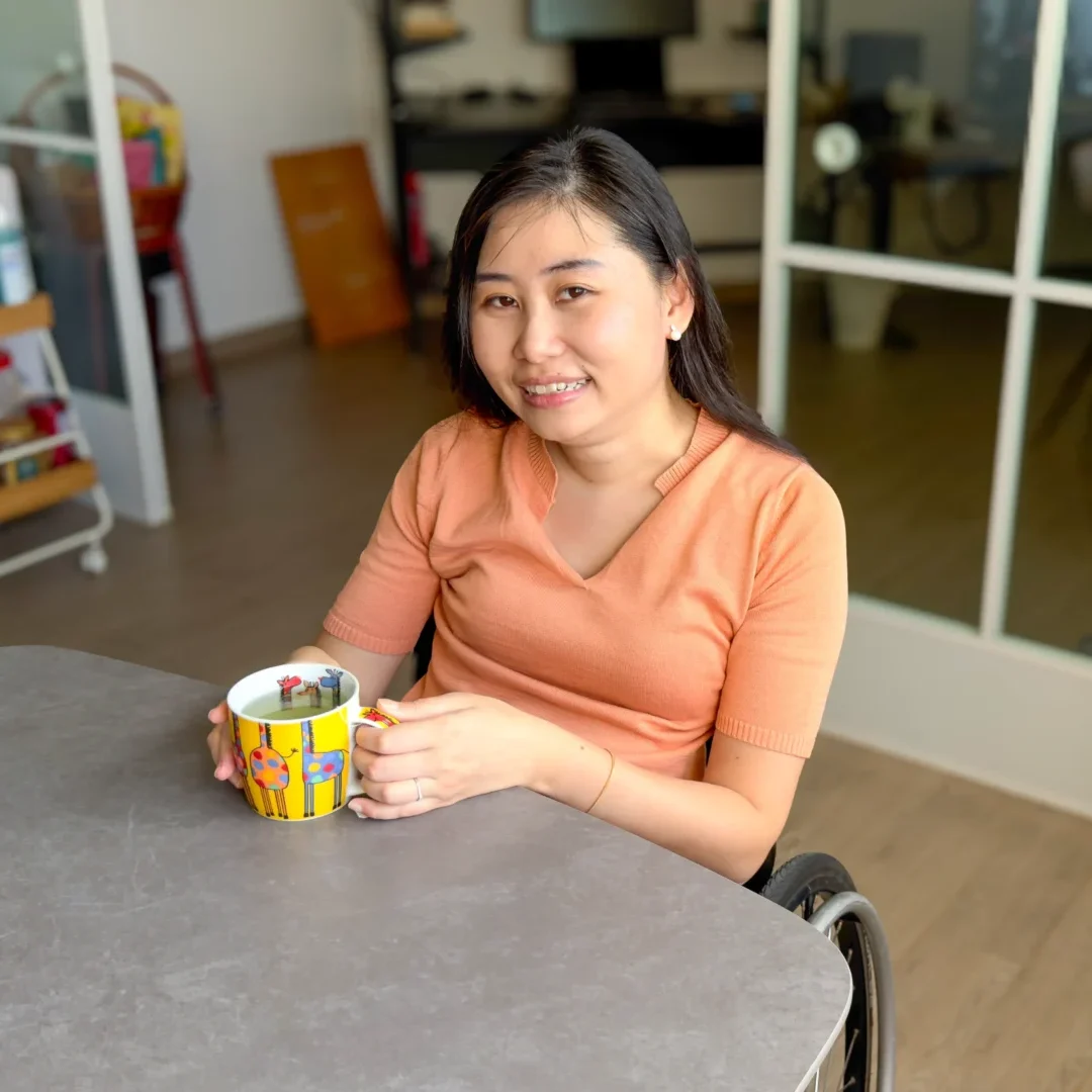 Amanda Chan, a young woman seated in her wheelchair, drinking a cup of tea at her dining table.