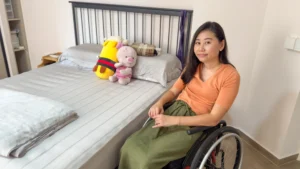 Amanda seated in her wheelchair, posing next to her bed. The bed is the same height as her wheelchair.