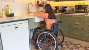 Amanda has her wheelchair pushed up to the kitchen sink, which has an open space underneath.