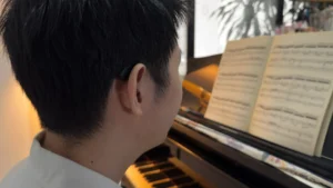 A closeup of Azariah’s hearings aids from the back. In front of him on the piano, there’s an open book of sheet music.