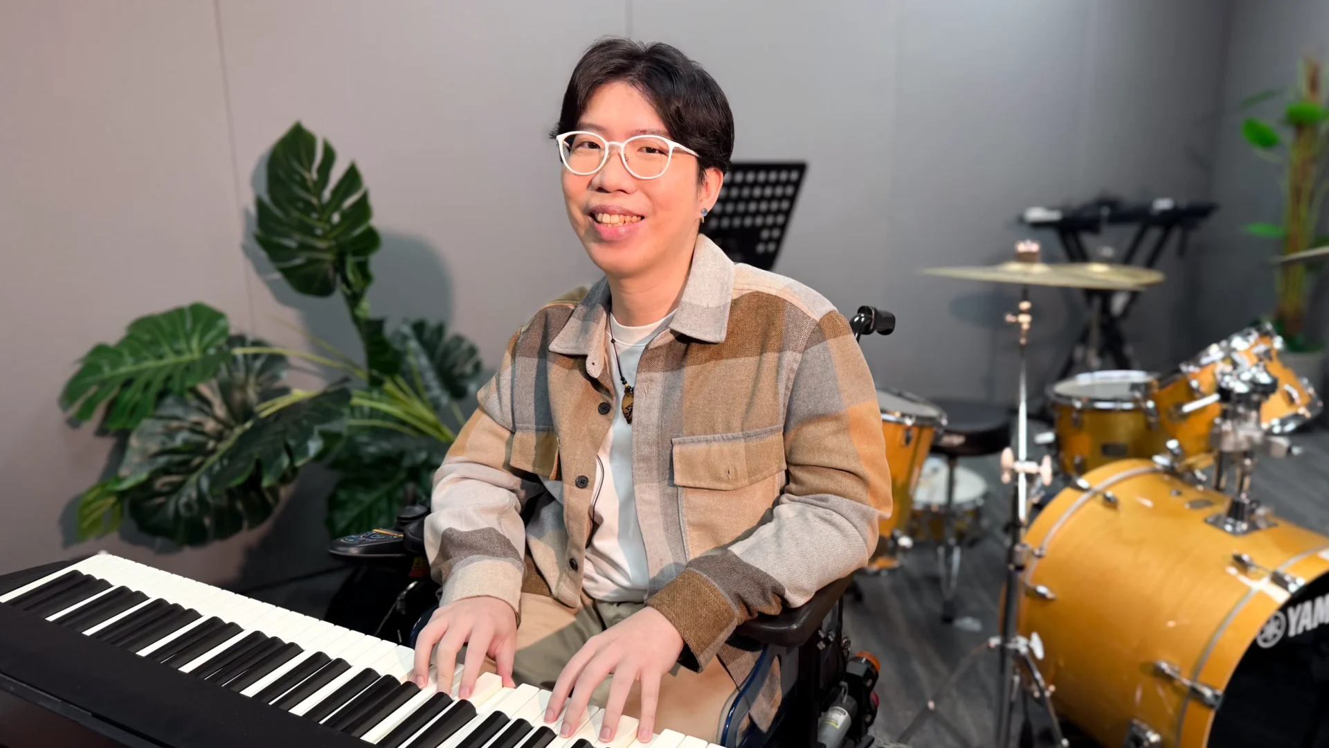 Sky Shen smiling at the camera, seated in his wheelchair. He is posing with an electric keyboard in a recording studio.