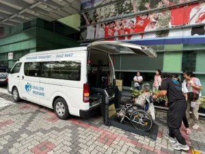 A woman in a wheelchair is entering a wheelchair transport van via the mechanical lift at the back of the vehicle. The name of the wheelchair transport business, Shalom Medcare, is printed on the side of the van.